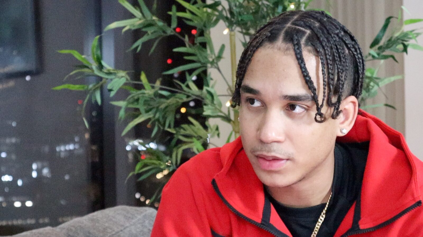 Joshua Arthur on camera in a red tracksuit, braids, a city window behind him